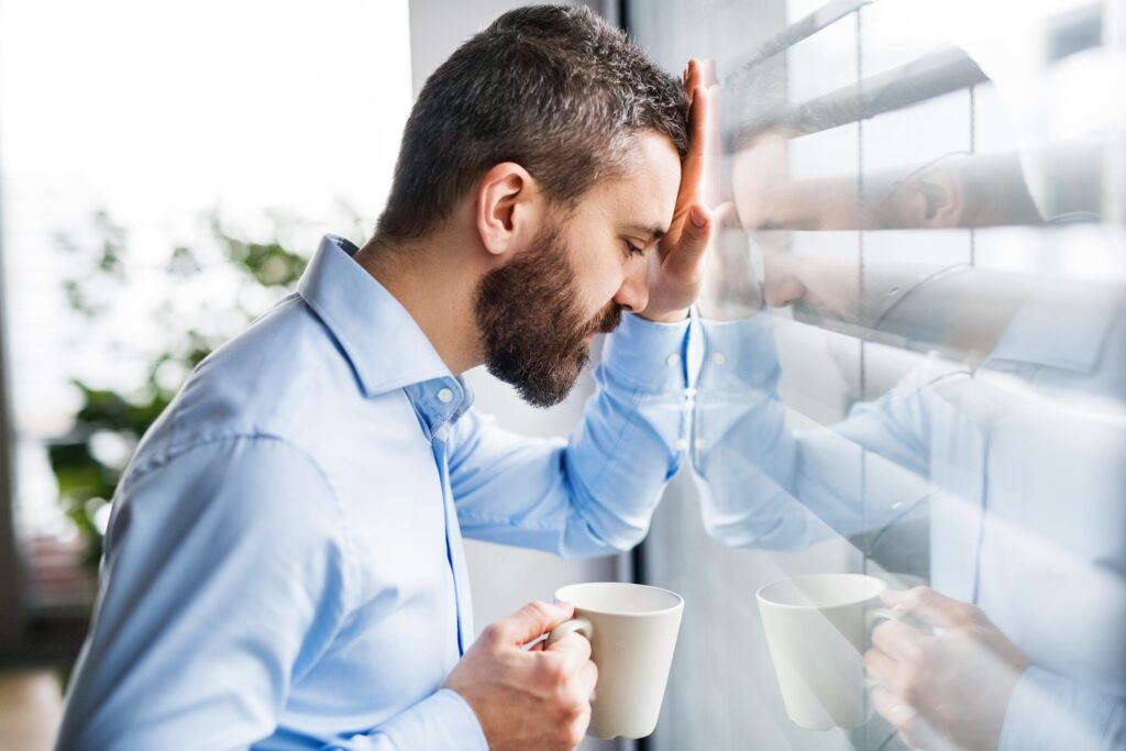 Frustrated office worker holding a mug and leaning his head against a window, clearly unhappy with his coffee.