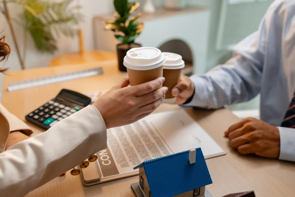 Estate agent offering coffee to a client during a property meeting, with documents, a calculator and a model house on the desk.